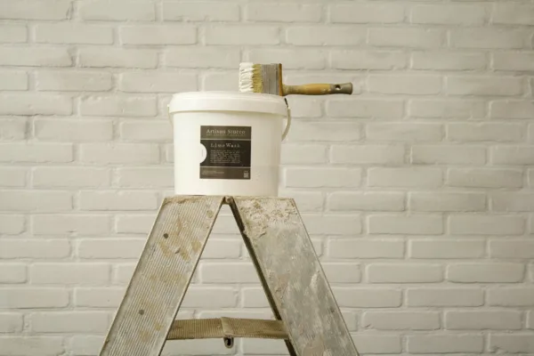 Artisan Stucco Ebro White Lime Wash paint bucket with brush on top of a ladder and with with painted stone wall in the background